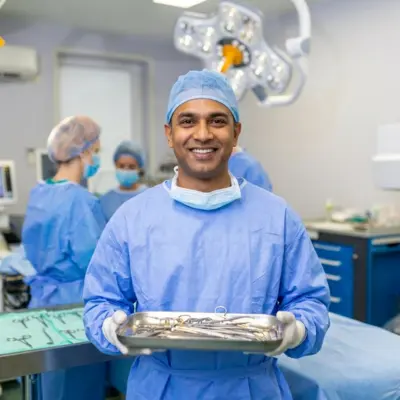 A male surgical technician smiling while holding a tray of instruments in an operating room