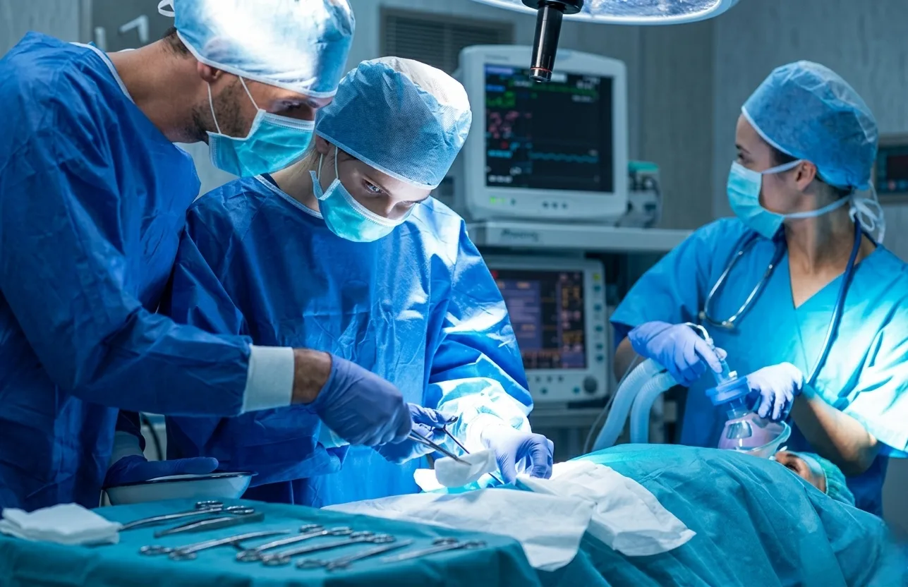 A surgical team in blue scrubs and masks performing a procedure under bright operating room lights.