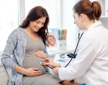 A female doctor uses a stethoscope to examine a smiling pregnant woman in a bright