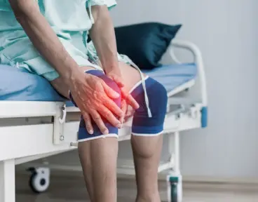 A patient in a hospital gown sitting on a bed, holding a painful knee highlighted with a red glow.