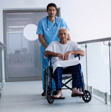 A male nurse in blue scrubs pushes an elderly patient in a wheelchair down a hospital hallway.