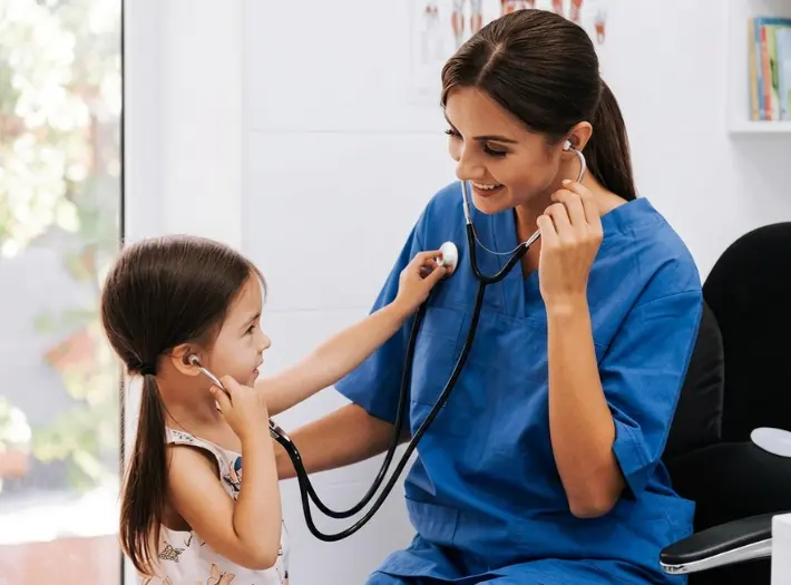 A female doctor in blue scrubs smiling as a young girl listens to her heartbeat with a stethoscope.