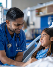 A male nurse in blue scrubs leans over a hospital bed to talk with a happy young girl.